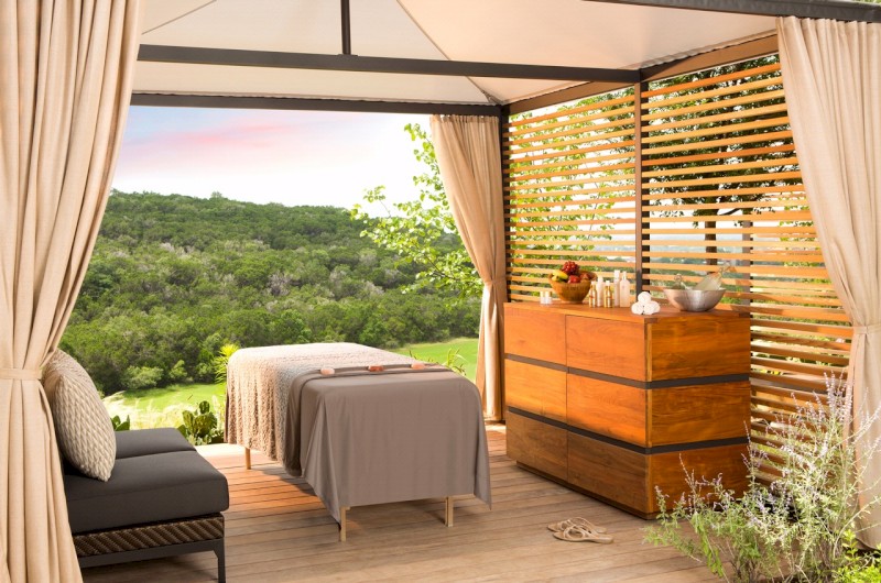 A peaceful, open-air room with a massage table, chest of drawers, and view of lush trees and greenery, creating a serene and relaxing atmosphere.